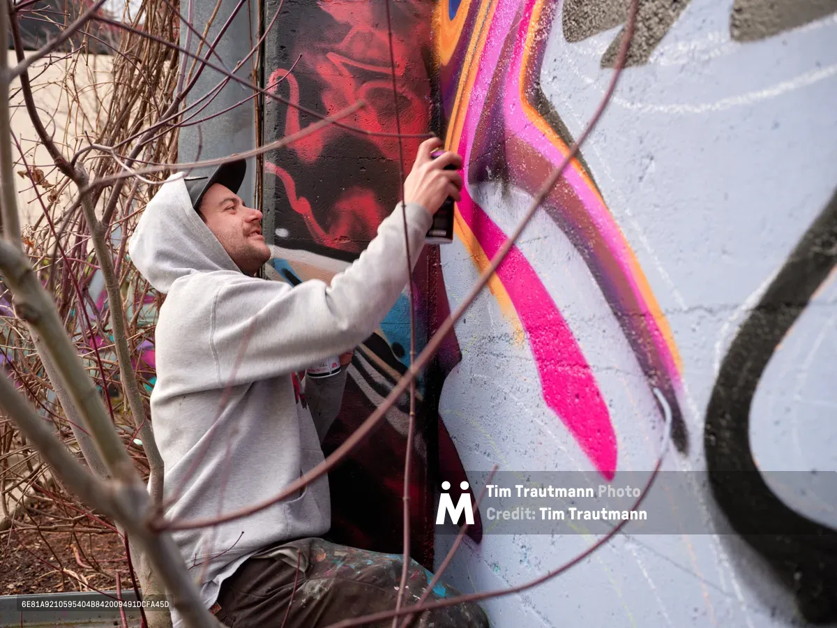 Travis Fields, known as Campographic, applies vibrant magenta and orange paint to a concrete wall at the Clay Creative Building in southeast Portland's Hosford-Abernethy neighborhood. Crouched behind protective metal fencing, the hooded artist methodically works on his mural while winter-bare branches frame the scene. The industrial setting and natural lighting create an authentic documentary atmosphere capturing the intersection of urban art and creative community spaces.