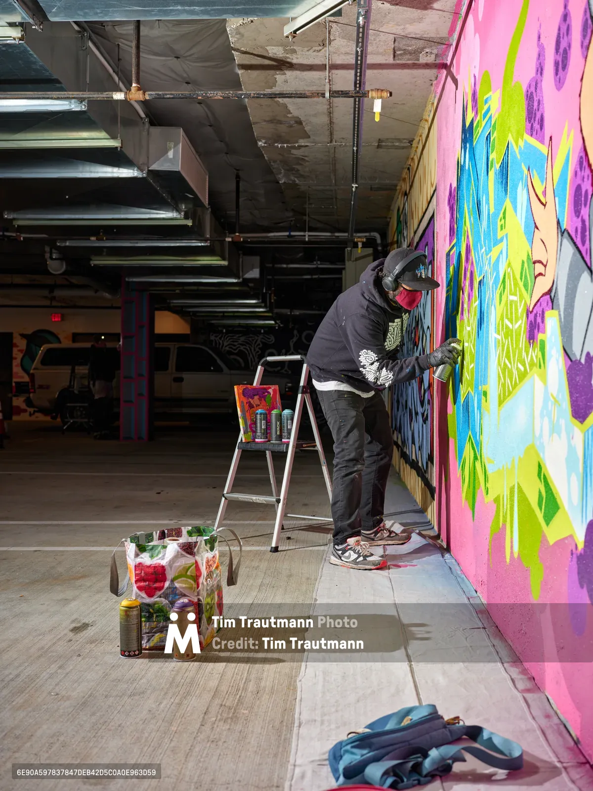 A hooded artist in dark clothing meticulously works on a large-scale mural featuring explosive bursts of pink, yellow, blue and purple paint across the concrete wall of a converted parking garage. The industrial space at Clay Creative in Southeast Portland's Hosford-Abernethy neighborhood buzzes with creative energy as exposed ceiling infrastructure frames the scene. Spray paint cans, brushes, and art supplies scatter across the polished concrete floor, while natural light filters through the urban warehouse setting, illuminating the transformation of raw space into artistic canvas.