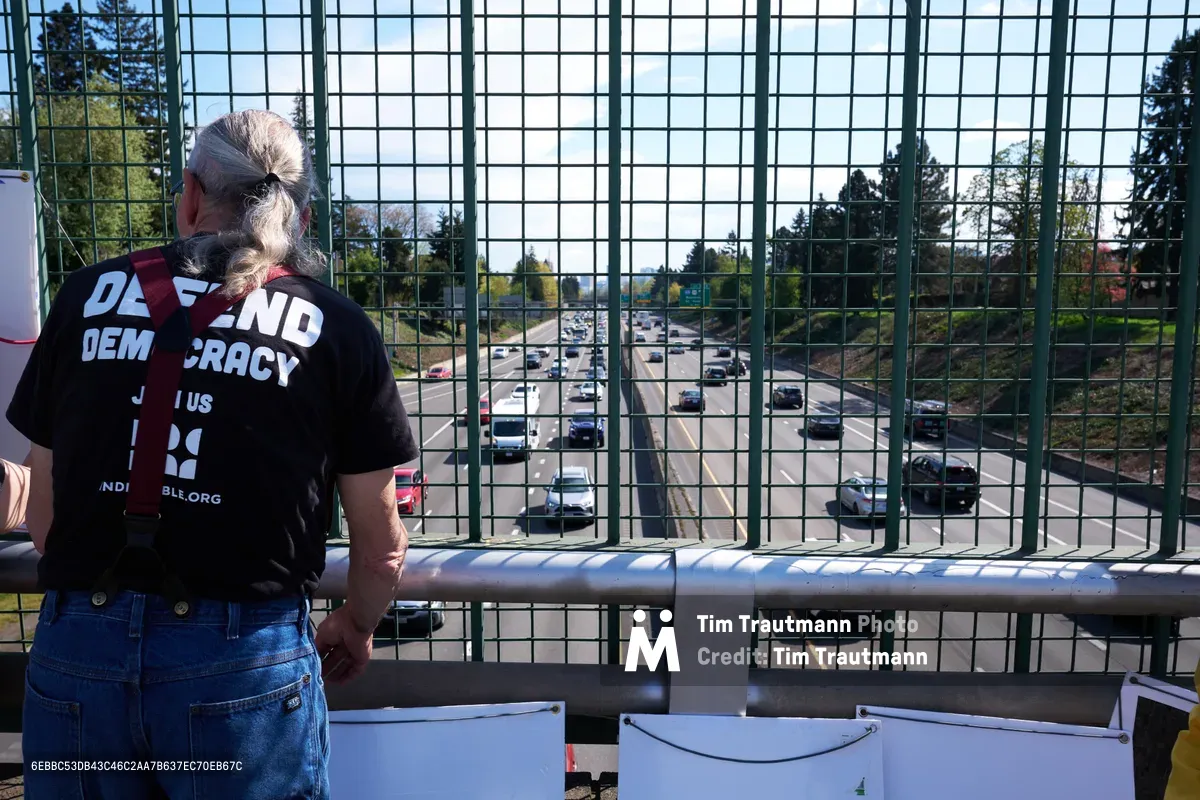 A silver-haired activist wearing a "Defend Democracy" t-shirt stands at a chain-link fence overlooking Interstate 5 in North Portland, participating in a coordinated banner drop action. The woman grips the metal railing as traffic streams below through the urban corridor, framed by evergreen trees and overcast Pacific Northwest skies. White banner materials are visible at the base of the overpass, ready for deployment in this grassroots political demonstration calling for a May 1st General Strike.
