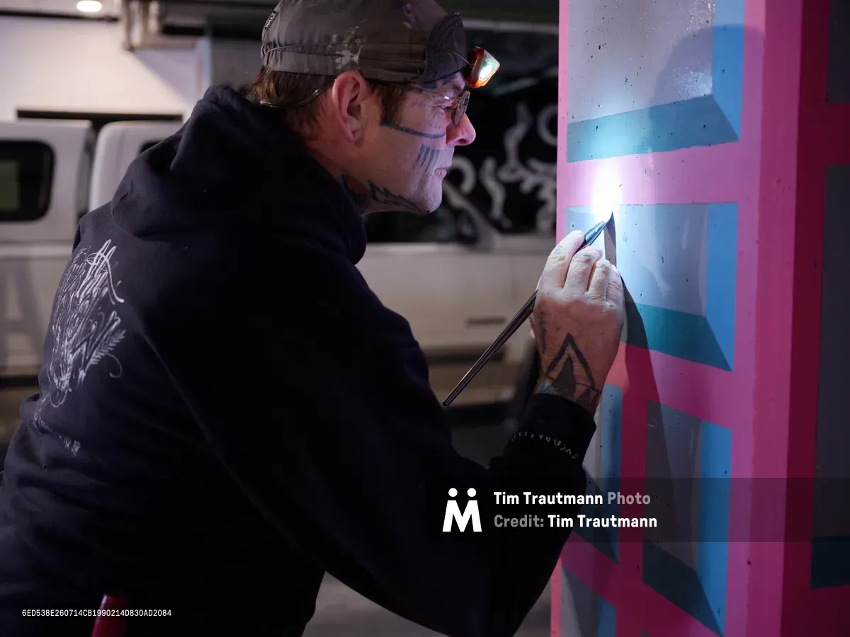 Artist Rabbitt meticulously applies precise brushstrokes to a vibrant geometric mural within the industrial confines of Portland's Clay Creative building parking garage. His tattooed hands work with steady concentration as magenta and cyan geometric forms emerge on the concrete wall, illuminated by a focused headlamp that cuts through the garage's dim atmosphere. The juxtaposition of raw urban infrastructure and carefully crafted geometric patterns captures the essence of Portland's underground art scene.