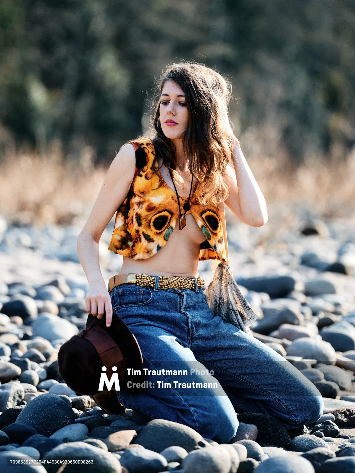 A young woman sits contemplatively among smooth river stones along Oregon's Floodplain Trail, her chestnut hair catching golden afternoon light. She wears a striking animal-print crop top paired with high-waisted denim and layered accessories, embodying free-spirited bohemian style. The shallow depth of field isolates her against the softly blurred forest backdrop, creating an intimate portrait that speaks to wanderlust and connection with nature.