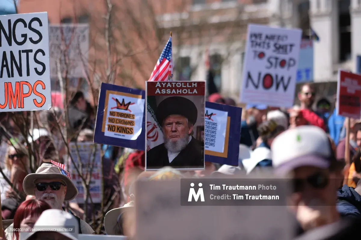 A determined crowd fills Southwest Ankeny Street in Portland's Old Town Historic District during a 2026 anti-monarchy protest, their handmade signs creating a forest of dissent against the brick facades of Chinatown. At the demonstration's heart, a provocative poster depicting a political figure as 'Ayatollah Assahola' towers above the sea of protesters, while American flags flutter defiantly beside placards declaring 'No Thrones No Crowns No Kings.' The afternoon light casts sharp shadows across the diverse gathering, capturing the raw energy of civic resistance in the Pacific Northwest.