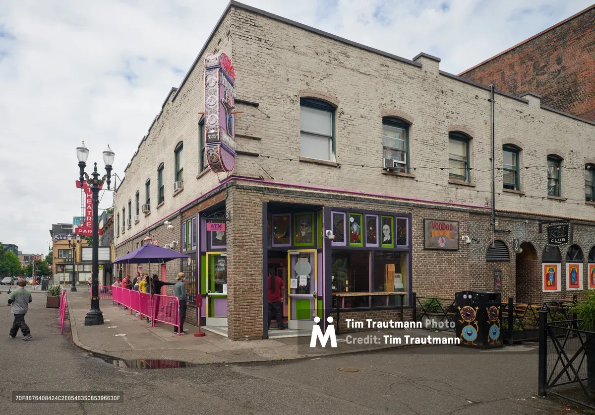 The iconic Voodoo Doughnut shop anchors a weathered brick corner in Portland's Old Town Historic District, its signature purple and green storefront radiating defiant vibrancy against overcast skies. Pink safety barriers channel a socially distanced queue of masked customers along the sidewalk, while the establishment's eclectic signage and window displays maintain their characteristic irreverent charm. The scene captures urban resilience during the pandemic, with pedestrians navigating the altered streetscape beneath heavy Pacific Northwest clouds.