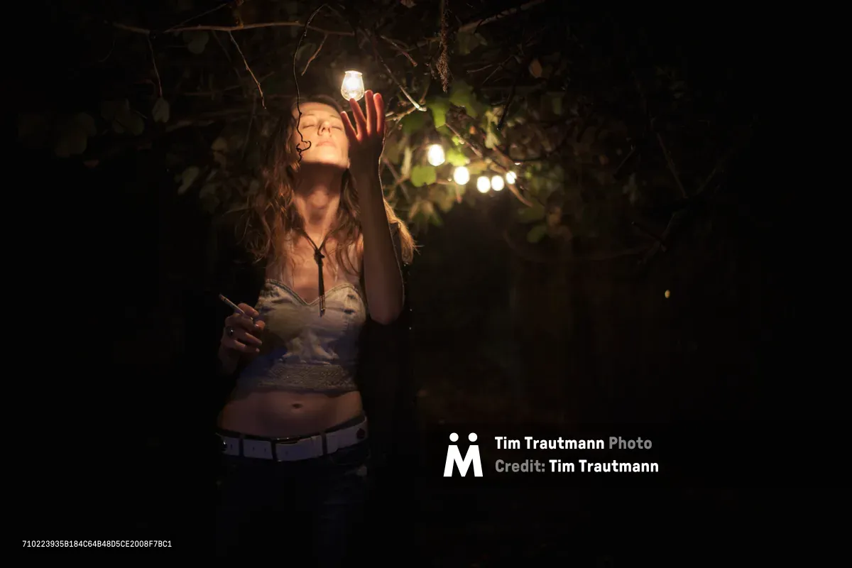 A young woman with flowing hair reaches upward toward a suspended light bulb in a darkened outdoor space in Portland's Humboldt neighborhood. String lights create a dreamy bokeh background among tangled branches and foliage, while her contemplative expression and graceful gesture embody a moment of connection with illumination. The atmospheric scene captures the interplay between artificial light and natural darkness in an intimate urban garden setting.