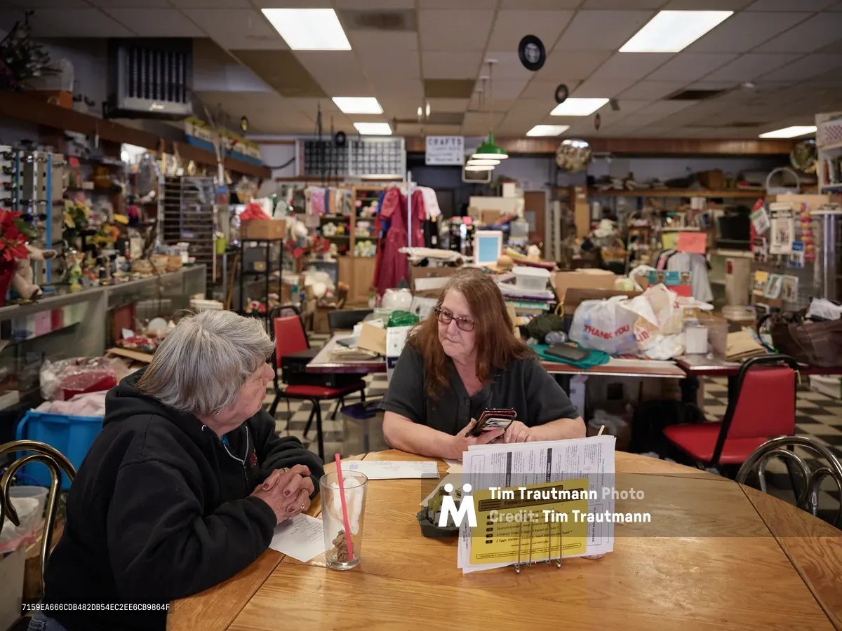 Pattie Deitz, owner of Pattie's Home Plate Café, sits across from her best friend Jenny at a wooden table in her eclectic North Portland restaurant. The warm fluorescent lighting illuminates scattered papers and a yellow menu holder between them, while the cluttered, homey interior filled with collectibles and memorabilia creates an intimate backdrop. Their relaxed conversation captures the intersection of friendship and small business ownership in the Saint Johns neighborhood.