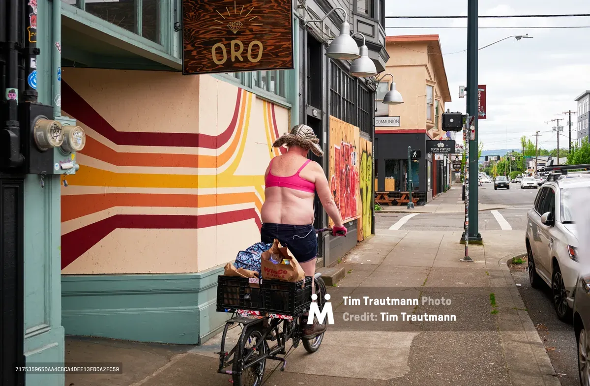 A woman in a bright pink bikini top and dark shorts cycles past her cargo bicycle on Southeast Hawthorne Boulevard in Portland, her back to the camera as she moves along a storefront adorned with vibrant geometric murals in warm sunset hues. The juxtaposition of her summer attire against the backdrop of early pandemic business closures captures a moment of personal freedom amid the uncertainty of COVID-19 lockdowns. Industrial pendant lights hang from the mint-green building facade, while parked cars and power lines stretch toward distant mountains under an overcast Pacific Northwest sky.