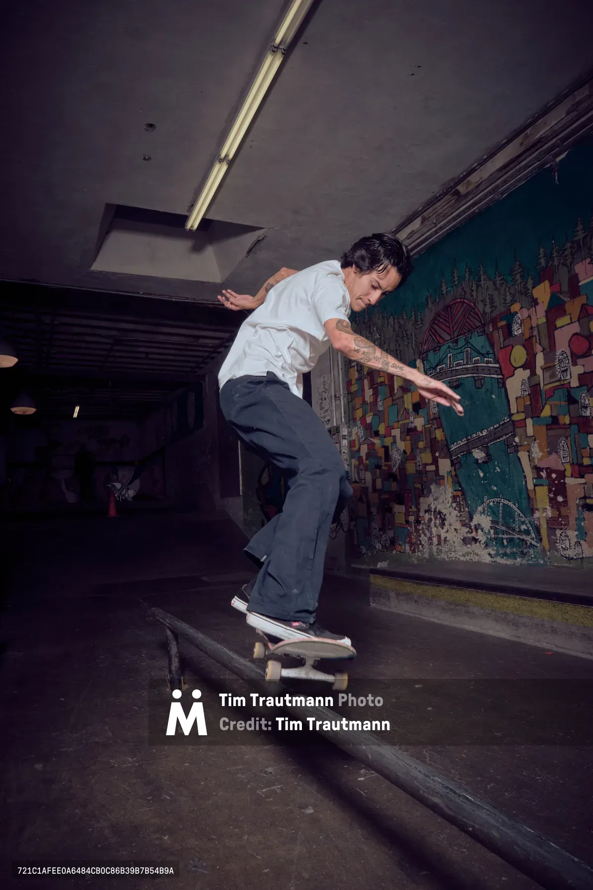 A skateboarder in crisp white shirt and dark pants executes a smooth rail grind in the atmospheric underground space of Commonwealth Skateboarding in Portland's Buckman neighborhood. The moody lighting creates dramatic shadows against weathered concrete walls adorned with vibrant street art murals, while industrial ceiling fixtures cast pools of golden light across the worn wooden rails. The scene captures the raw authenticity of Portland's skateboarding culture within this iconic Southeast 20th Avenue skate shop.