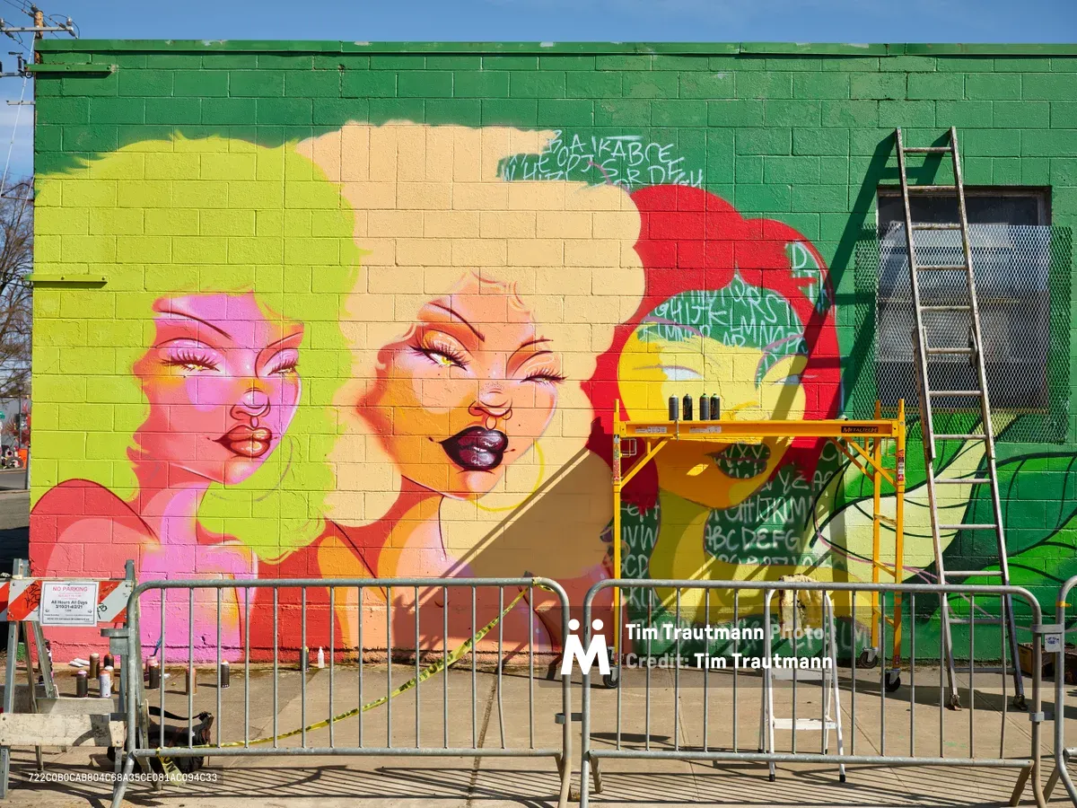 A striking work-in-progress mural titled Jardín Cubana dominates a lime green cinderblock wall along Northeast Alberta Street in Portland's Alberta Arts District. The large-scale artwork features three glamorous female figures rendered in vivid pinks, yellows, and oranges, their faces emerging from swirling colorful hair against the emerald backdrop. Construction scaffolding and safety barriers frame the scene, capturing multidisciplinary artist Vilatose's creative process in this gentrifying neighborhood known for its street art culture.