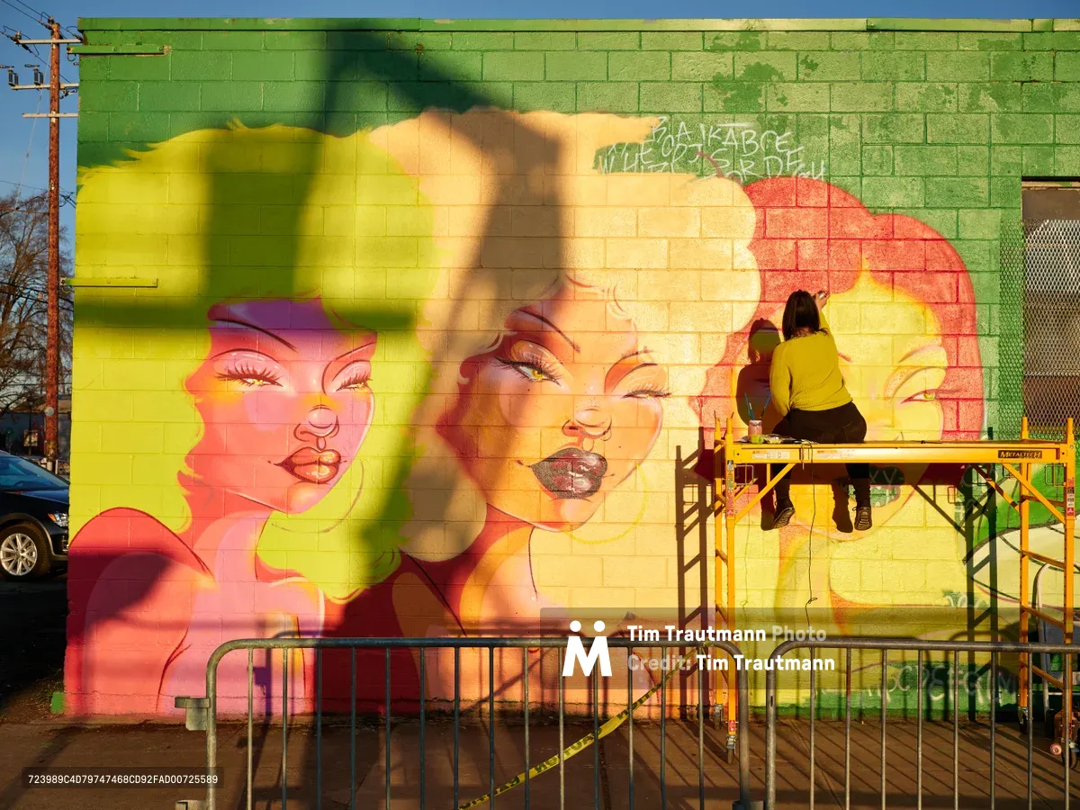 Multidisciplinary artist Vilatose works from a yellow scaffolding platform, adding finishing touches to 'Jardín Cubana,' a striking mural featuring three female figures rendered in brilliant coral, yellow, and green hues on a cinder block wall. The large-scale artwork depicts stylized feminine portraits with flowing hair, painted across the green brick facade in Portland's Alberta Arts District. Golden hour light bathes the scene, casting warm shadows while the artist concentrates on their craft, surrounded by construction barriers and urban infrastructure.