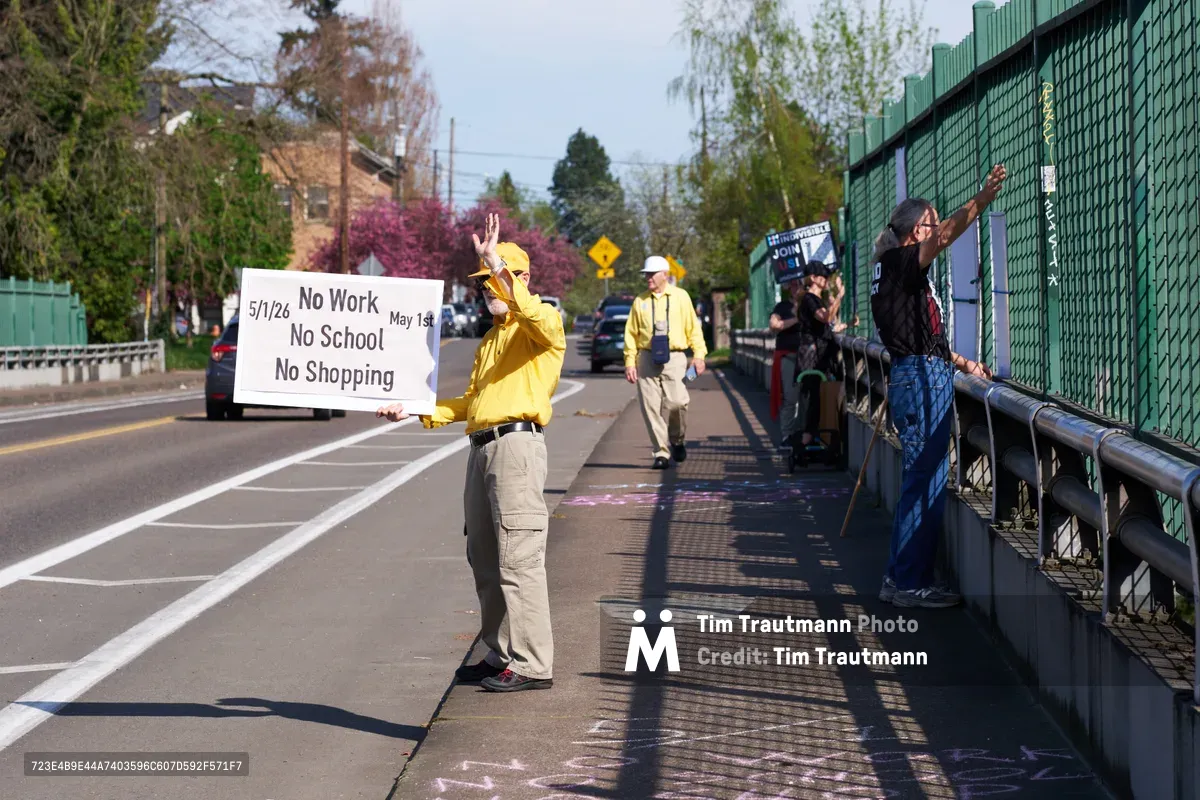 Activists in bright yellow rain gear hold protest signs on North Skidmore Street bridge over Interstate 5 in North Portland during a May Day banner drop action. The central figure displays a white banner reading "No Work No School No Shopping May 1st" while others raise their arms in solidarity. Spring foliage frames the scene as cars pass beneath the overpass, creating a moment of grassroots political theater against the suburban Portland backdrop.