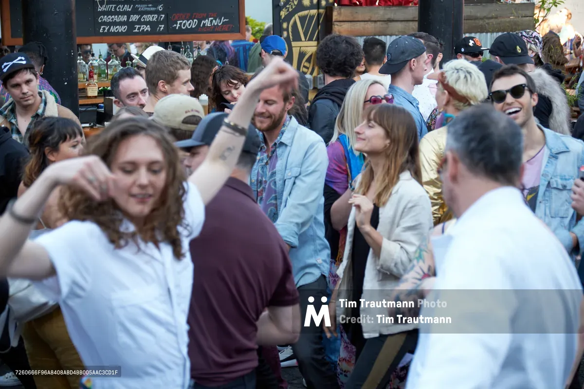 A vibrant crowd of young adults mingles in the warm afternoon light at White Owl Social Club during the Sunday Best season opener, a popular summer day party in Portland, Oregon. The diverse gathering shows people in casual summer attire—denim shirts, baseball caps, and sunglasses—creating an atmosphere of relaxed social connection. Behind the animated conversations, chalkboard menus advertising craft beverages hint at the venue's artisanal character. The scene captures the infectious energy of Portland's music and social scene as friends reconnect and new acquaintances form in the dappled sunlight.