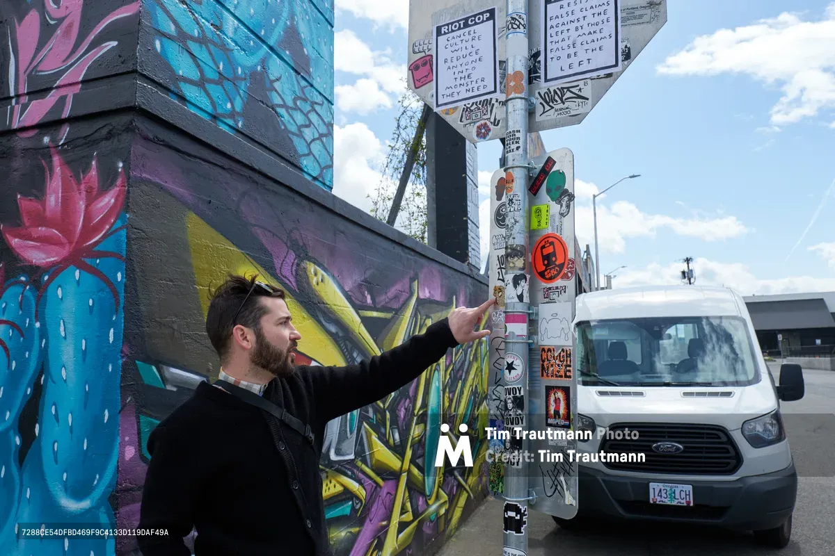 Tomás Valladares of the Portland Street Art Alliance gestures toward a utility pole densely layered with stickers and tags during a guided tour in the Hosford-Abernethy district. The scene unfolds against vibrant graffiti murals featuring blue and pink floral motifs, while a white Ford Transit van waits nearby under an overcast afternoon sky. His animated explanation captures the intersection of grassroots artistic expression and urban culture documentation in Portland's creative corridors.