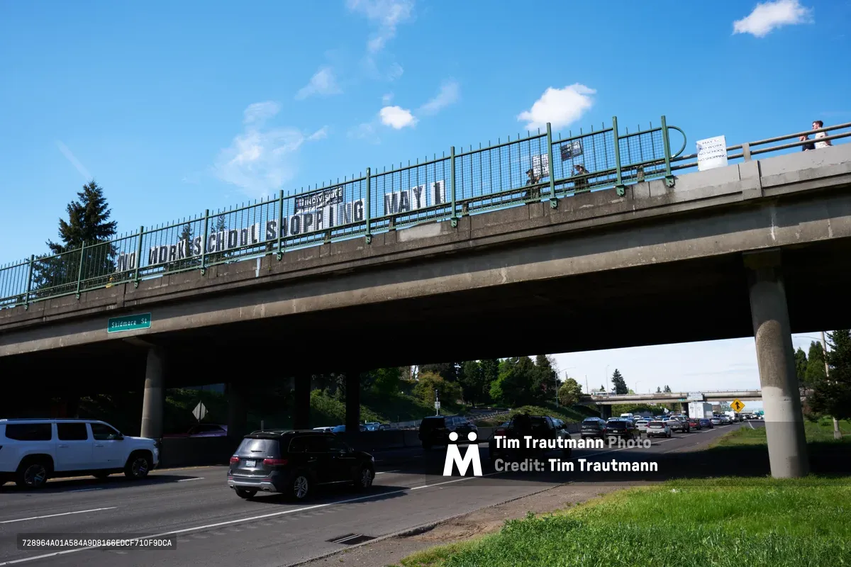 Activists from the Indivisible movement unfurl a protest banner across the Interstate 5 overpass at North Skidmore Street in Portland, Oregon, calling for a May 1st general strike. The white banner with bold black lettering reading 'NO WORK SCHOOL SHOPPING MAY 1st' spans the concrete bridge structure above busy freeway traffic. Against a brilliant blue sky dotted with wispy clouds, the grassroots demonstration transforms mundane urban infrastructure into a platform for political messaging, while commuter traffic flows beneath in the shadowed canyon of the highway corridor.