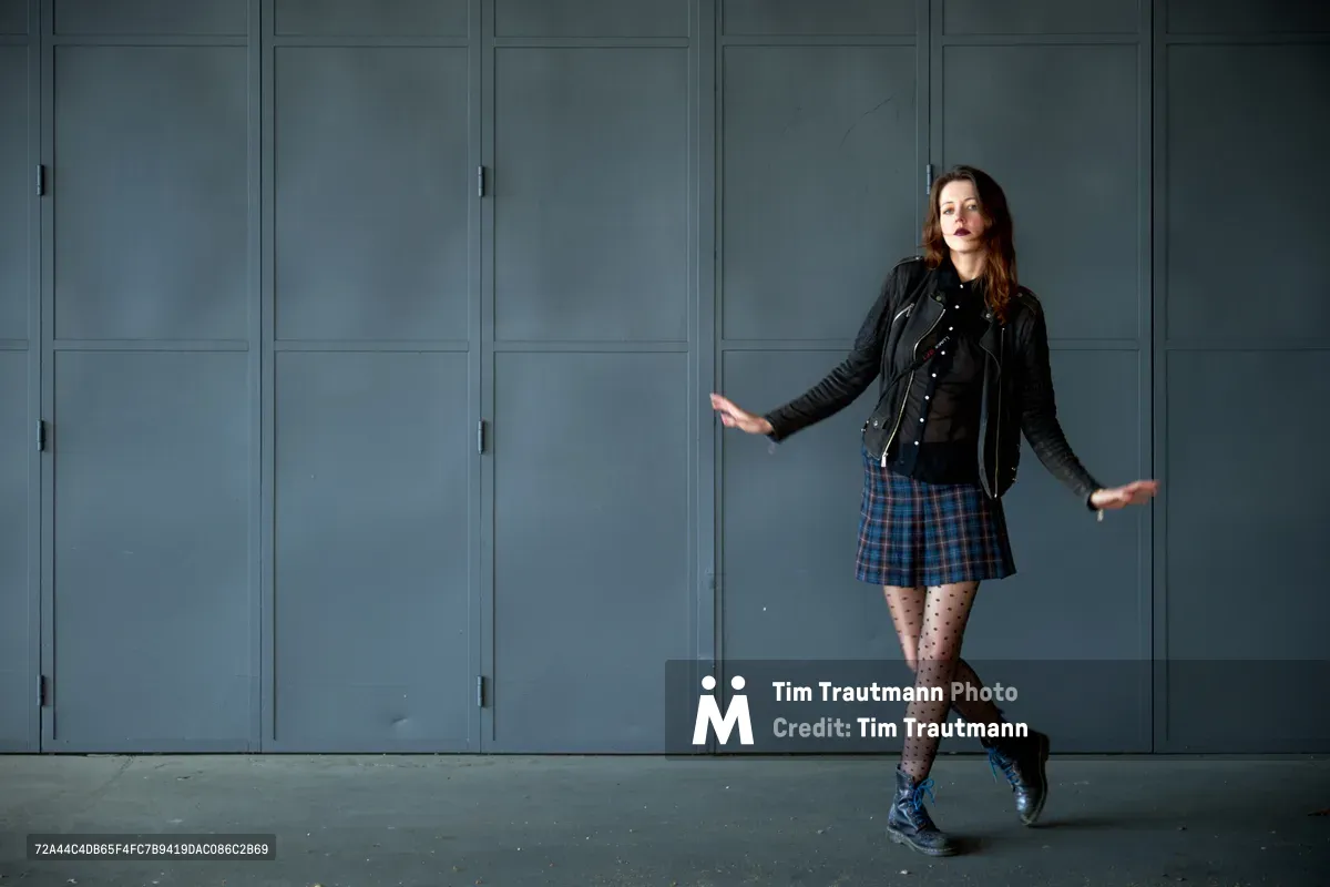 Rhianna Atwood strikes a dynamic pose against the steel blue industrial lockers of Portland's Central Eastside district. Her edgy ensemble of black leather jacket, plaid skirt, and polka dot tights creates a striking contrast against the muted urban backdrop. The natural lighting casts subtle shadows while capturing the confident energy of this editorial test shoot in the industrial heart of Southeast Portland.