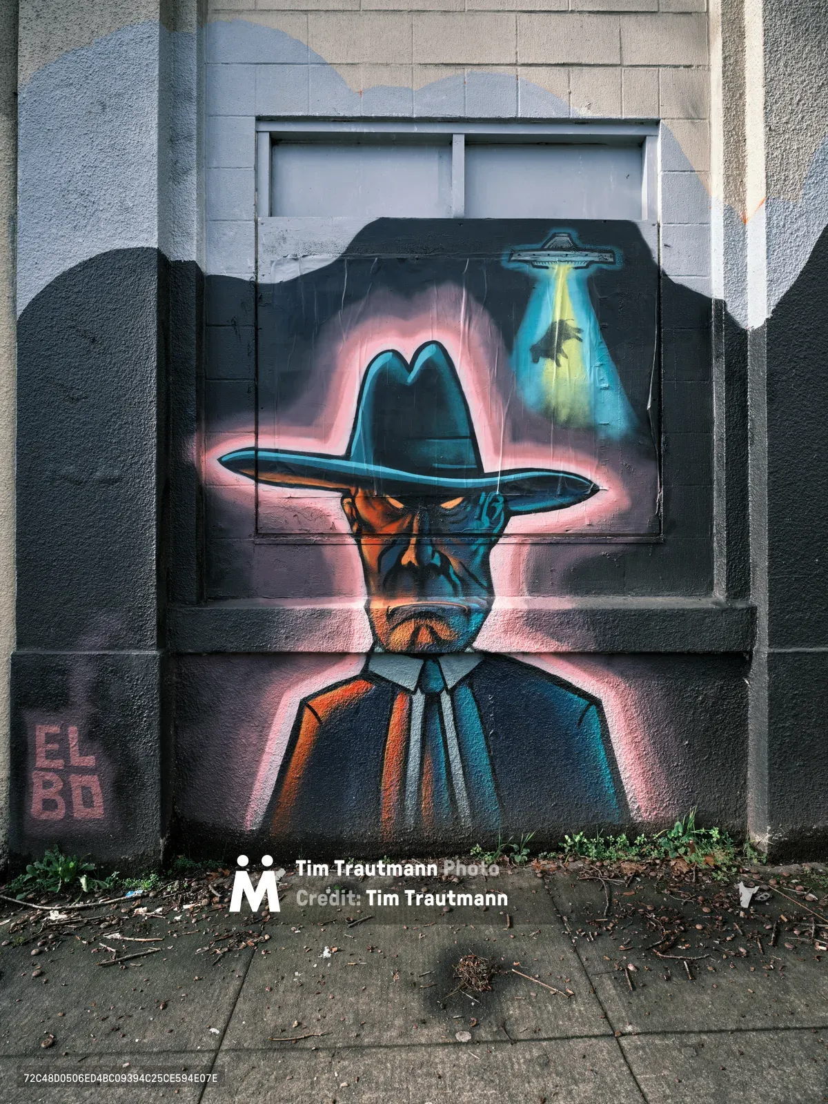 A striking street mural dominates the weathered concrete wall of the former Salvation Army warehouse in Portland's Central Eastside district. The artwork depicts a mysterious figure in a wide-brimmed cowboy hat and suit, rendered in dramatic blues and oranges with a glowing pink aura. Above the figure, a UFO casts an otherworldly teal beam of light, creating a surreal narrative of extraterrestrial encounter. The mural spans the lower portion of the industrial building, with cracked pavement and urban debris at its base, embodying the gritty aesthetic of Portland's commissioned street art scene.
