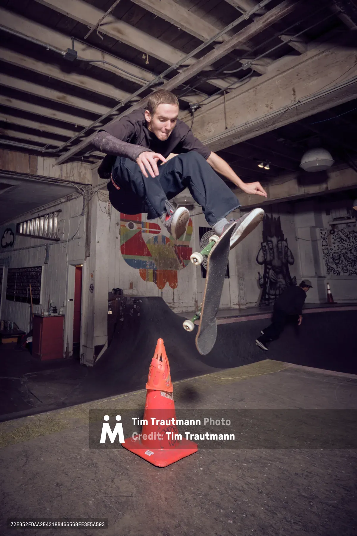 A young skateboarder launches into mid-air above a weathered orange traffic cone at Commonwealth Skateboarding in Portland's Buckman neighborhood. The exposed ceiling beams and graffiti-covered concrete walls create a raw, underground atmosphere, while another skater practices in the shadowed background. Dramatic lighting captures the precise moment of athletic suspension, highlighting the urban skate culture thriving beneath the city streets.