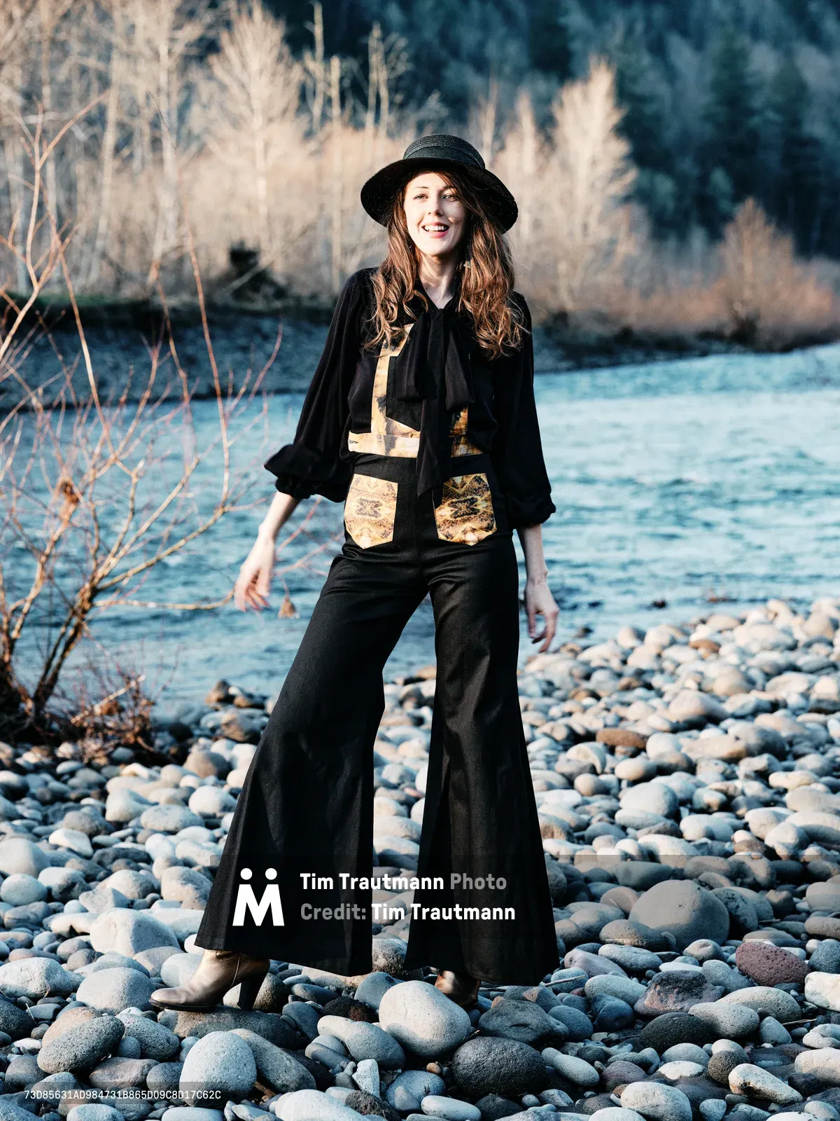A joyful woman in bohemian attire stands confidently on a pebbled riverbank along the Sandy River in Multnomah County, Oregon. She wears a black wide-brimmed hat, flowing black pants, and a distinctive vest with golden patches over a dark blouse. The late afternoon light illuminates her auburn hair and genuine smile, while dormant willows frame the rushing water behind her. The rocky riverbed stretches endlessly, capturing the serene wilderness of the Pacific Northwest.