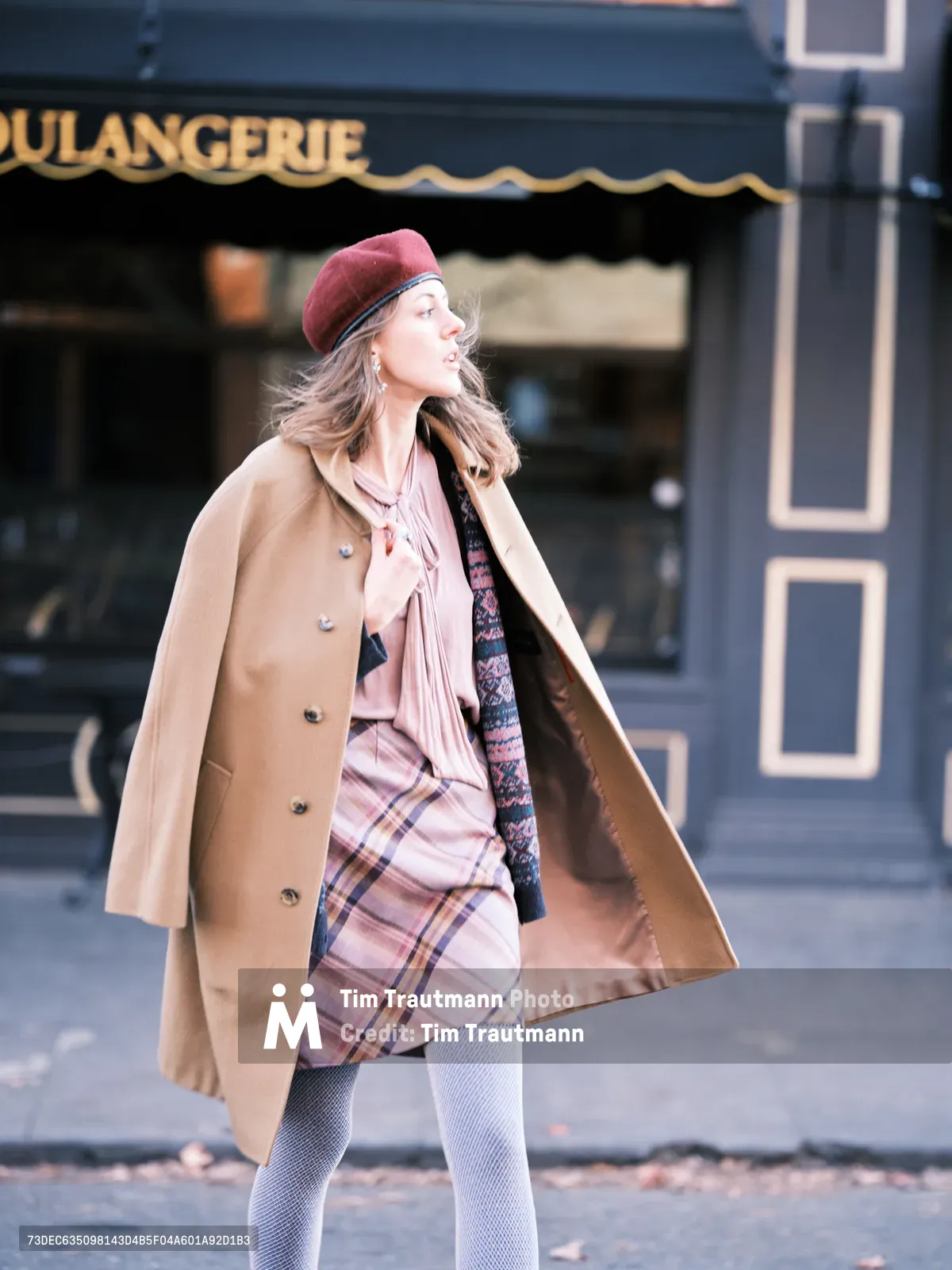 A stylishly dressed woman strides past a French boulangerie on Portland's Alberta Street, her burgundy beret and flowing camel coat catching the soft afternoon light. The layered autumn ensemble features a pink plaid skirt, blush scarf, and patterned tights, creating an effortlessly chic silhouette against the blue-gray storefront. Her hair flows behind her as she walks, embodying the relaxed sophistication of Northeast Portland's artistic corridor.