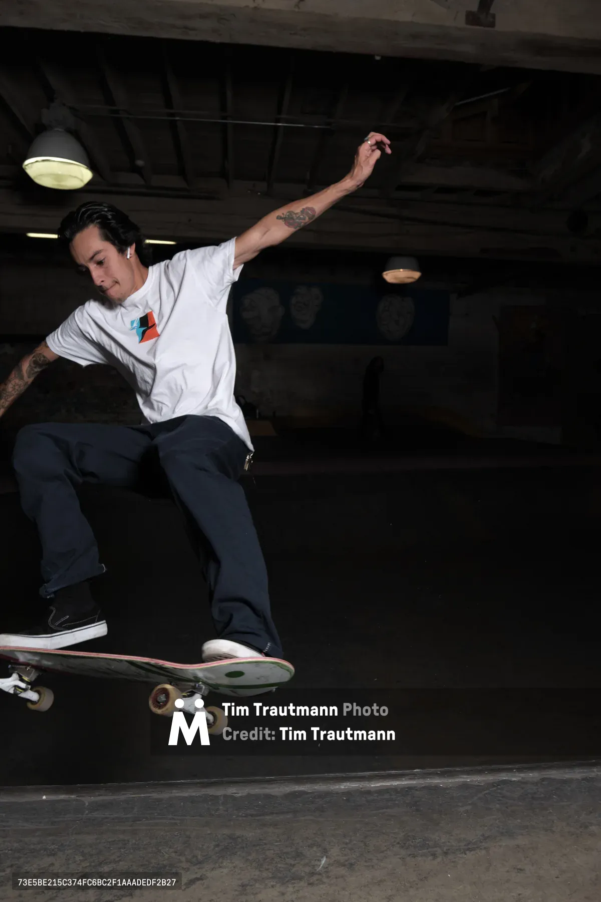 A tattooed skateboarder captures a moment of weightless artistry beneath the industrial ceiling of Commonwealth Skateboarding in Portland's Buckman neighborhood. His extended arm and focused expression reveal the meditative concentration required for technical skateboarding, while the harsh fluorescent lighting creates dramatic shadows against the concrete surfaces. The gritty basement atmosphere of the historic skate shop frames this editorial portrait of urban athleticism and creative expression.