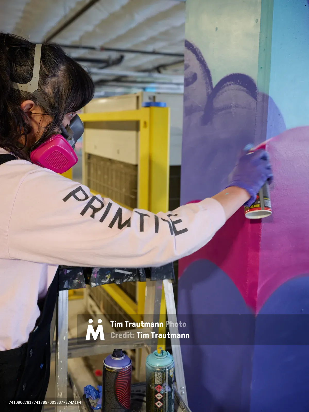 A muralist wearing a pink respirator mask and white sweatshirt emblazoned with 'PRIMITIVE' works on a vibrant purple and magenta mural in the industrial setting of Clay Creative's parking garage in Southeast Portland. Purple latex gloves protect her hands as she wields spray paint cans, their colorful caps arranged on a wooden cart below. The unfinished concrete ceiling and exposed infrastructure frame this creative transformation of utilitarian space into artistic expression.