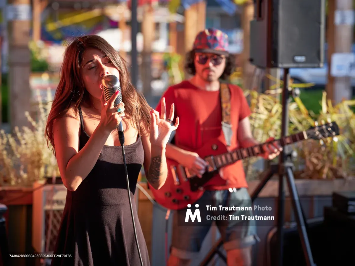 Vocalist Aubrey Gordon commands the stage with passionate intensity as golden late-afternoon light bathes her performance at Portland Mercado in Southeast Portland. Her expressive gestures and focused delivery create an intimate connection with the audience while her guitarist provides melodic accompaniment in the softly blurred background. The warm sunlight transforms the outdoor venue into a luminous stage, capturing the raw energy and authenticity of live street-side music in Oregon's vibrant cultural marketplace.