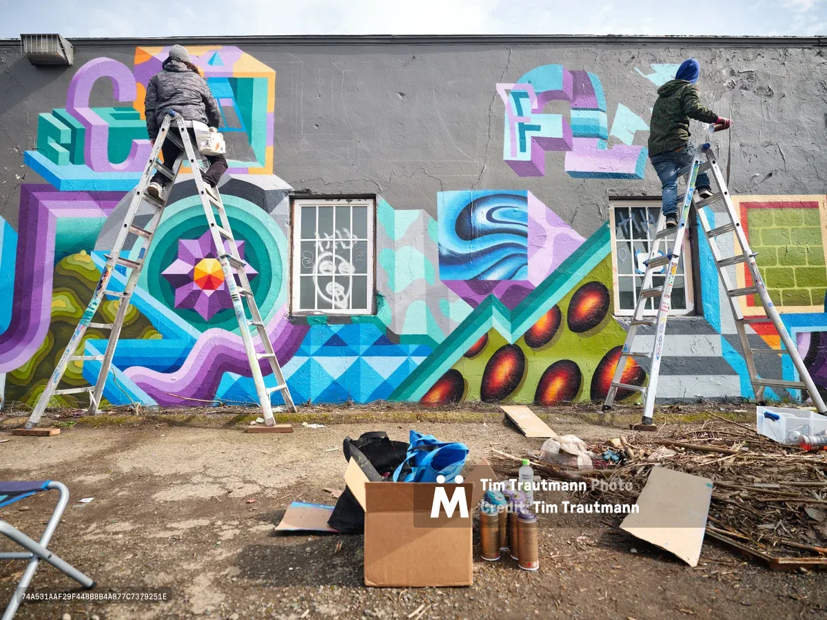 Two bundled-up street artists perch on aluminum ladders against a concrete building facade in Northeast Portland, Oregon, applying vibrant geometric patterns and dimensional lettering to create an expansive mural. The scene captures the collaborative process of urban art creation, with spray paint cans, cardboard supplies, and painting materials scattered across the debris-strewn ground below. Kaleidoscopic purples, teals, and oranges emerge from the weathered gray surface, while barred windows punctuate the artistic canvas under an overcast Pacific Northwest sky.