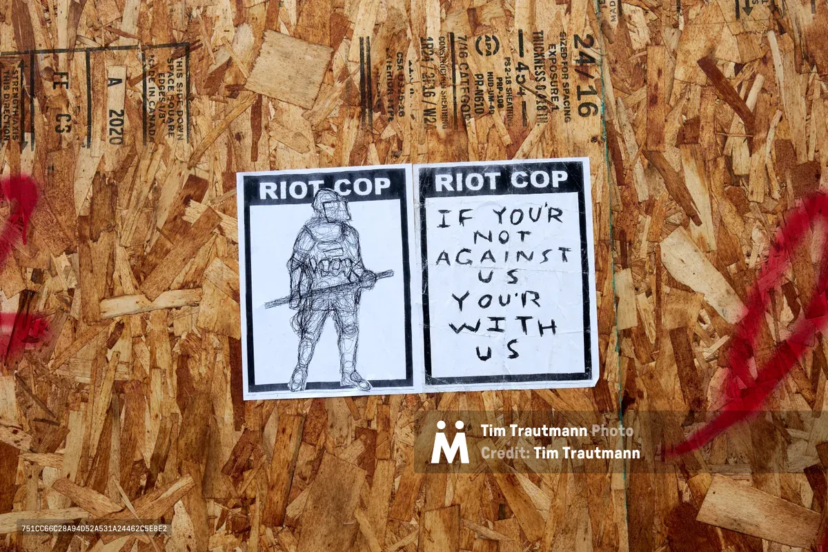 Two stark black and white stickers bearing "RIOT COP" headlines are affixed to weathered particle board covering a storefront window. The left sticker features a sketched figure of an armored officer, while the right declares "IF YOUR NOT AGAINST US YOUR WITH US" in hand-lettered text. Red spray paint marks streak across the textured wood surface, creating a raw urban canvas that speaks to Portland's turbulent protest era.