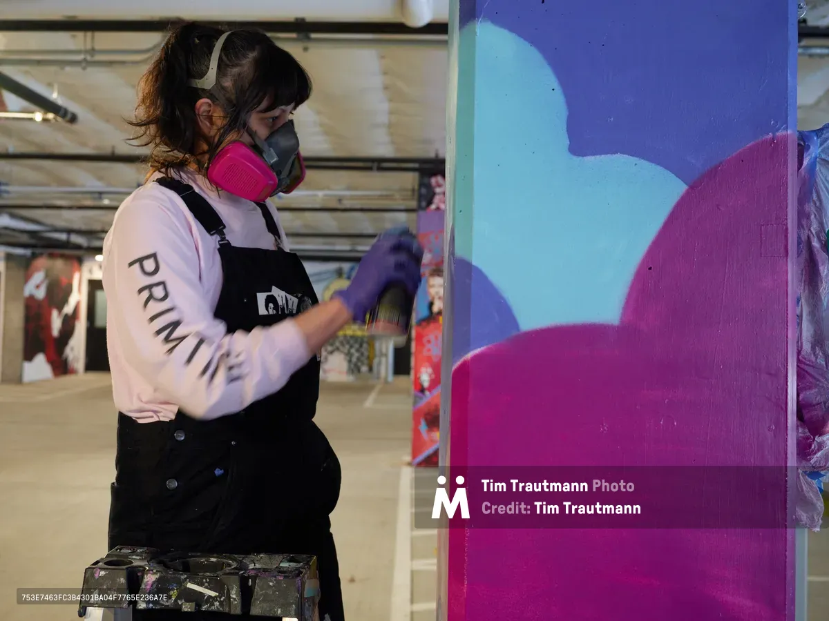 A focused muralist wearing a protective respirator and paint-splattered overalls applies bold magenta and turquoise paint to a large-scale wall piece in the industrial confines of Clay Creative's parking garage. The artist's concentrated brushwork reveals emerging abstract forms against the raw concrete backdrop, while scattered canvases and urban art installations populate the converted creative space in Southeast Portland's Hosford-Abernethy neighborhood.