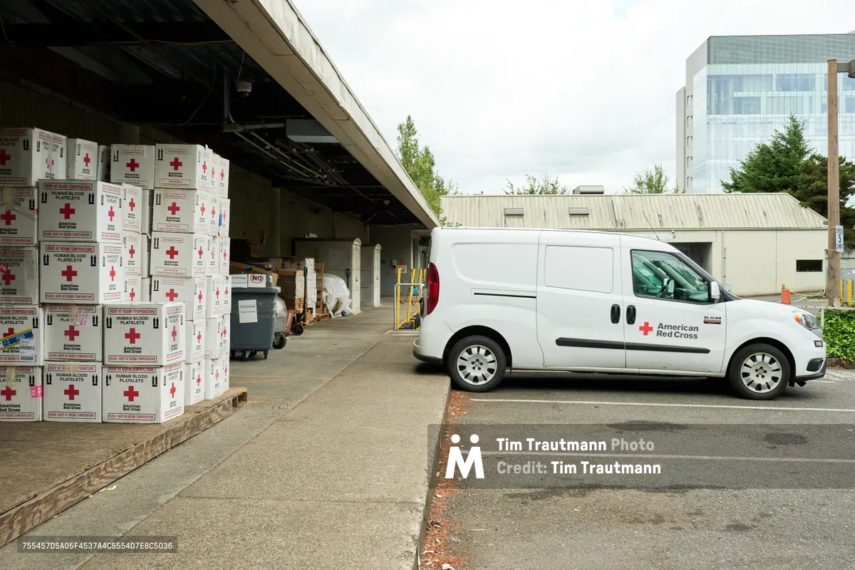 Under the industrial canopy of the American Red Cross facility in Portland's Albina neighborhood, towering stacks of empty human blood platelet boxes await their vital mission. A white delivery van bearing the iconic red cross emblem sits ready in the loading bay, while the urban backdrop of glass towers and overcast Pacific Northwest sky frames this essential humanitarian operation. The scene captures the quiet efficiency of medical supply distribution, where cardboard containers represent countless lives potentially saved through blood donation programs.