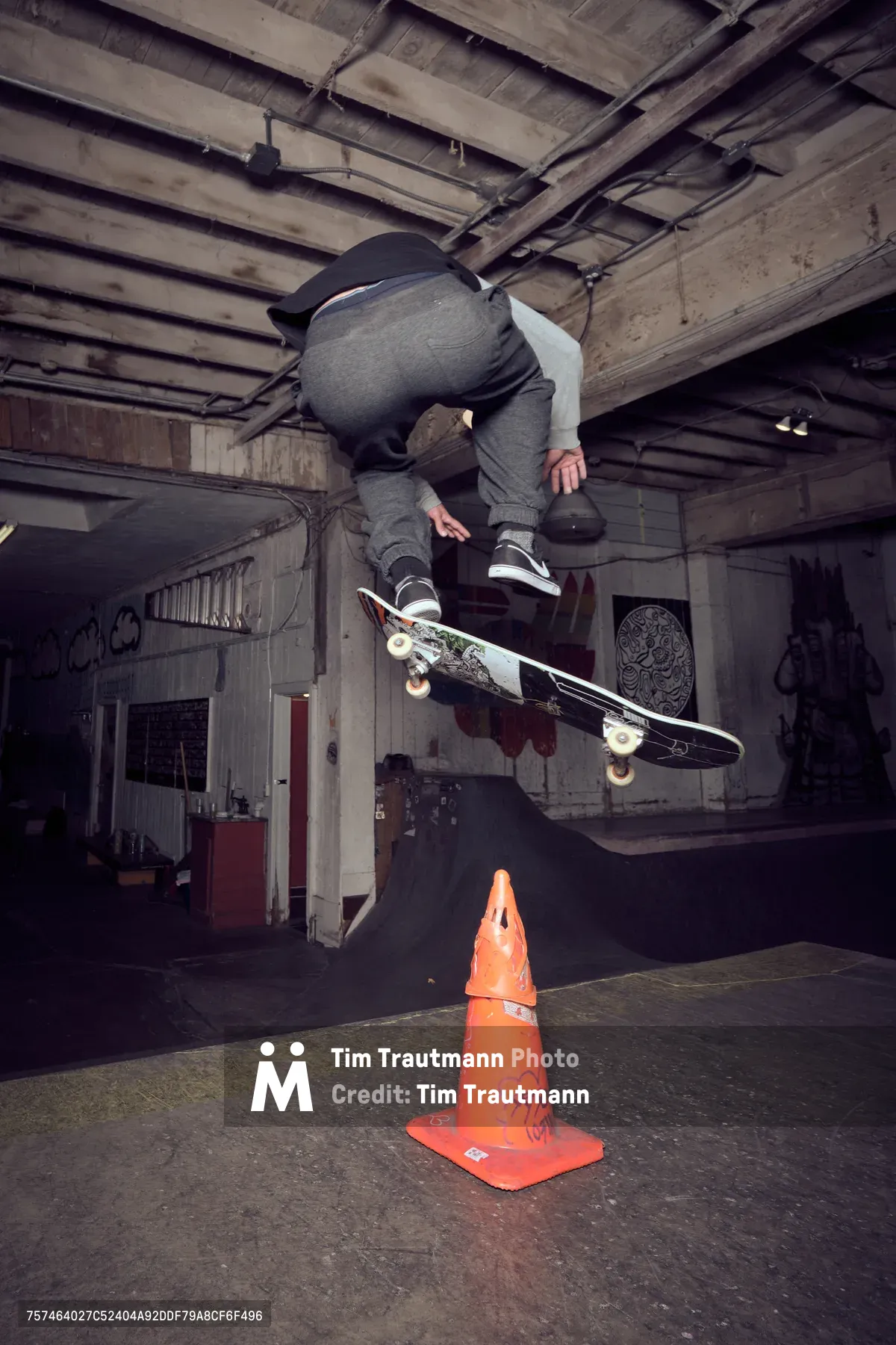 A skateboarder launches into a mid-air trick above a weathered orange traffic cone in the raw, industrial interior of Commonwealth Skateboarding in Southeast Portland. The basement space reveals exposed wooden ceiling joists and concrete walls adorned with graffiti, while dramatic shadows cast by overhead lighting create a gritty, underground atmosphere. The athlete, dressed in gray hoodie and dark jeans, demonstrates precise board control as his skateboard floats beneath him in this moment of athletic suspension.