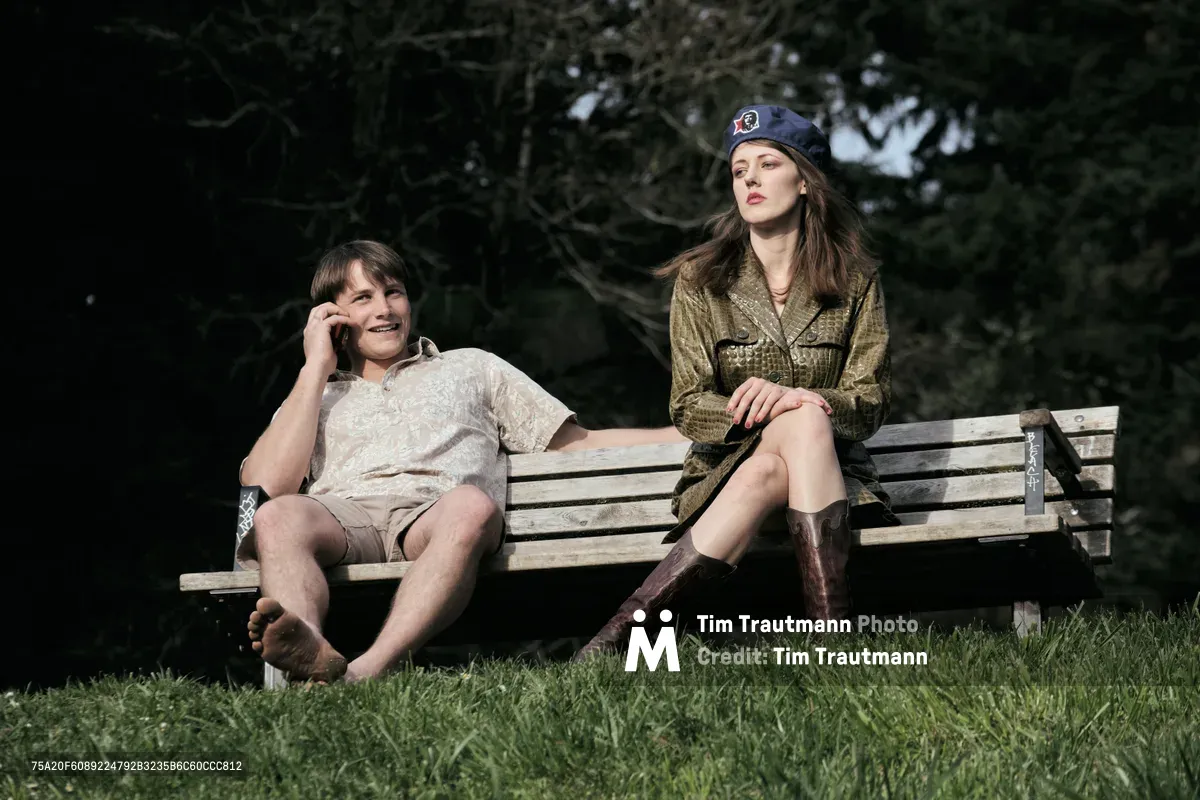 A young woman and young man share a quiet moment on a weathered wooden bench in Mount Tabor Park, Portland. The man on the left speaks into his phone while wearing casual shorts and summer attire, his companion sits poised in military-inspired olive green styling topped with a navy beret. Dappled afternoon light filters through the dense canopy above, casting gentle shadows across the emerald grass beneath their feet.