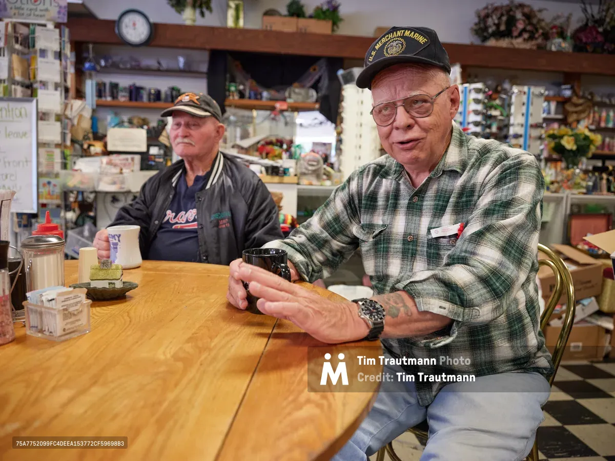 Two elderly regulars, Jewel and Ron, share stories at their corner table in Pattie's Home Plate Café in Portland's Saint Johns neighborhood. Ron, wearing a U.S. Merchant Marine cap and plaid flannel, gestures animatedly while speaking, his weathered hands conveying decades of experience. Jewel sits quietly beside him in a dark jacket, both men surrounded by the warm, cluttered atmosphere of their beloved local diner with its wood shelving lined with knickknacks and memorabilia. The golden wood table between them holds simple white mugs and condiments, testament to countless morning conversations in this community gathering place.