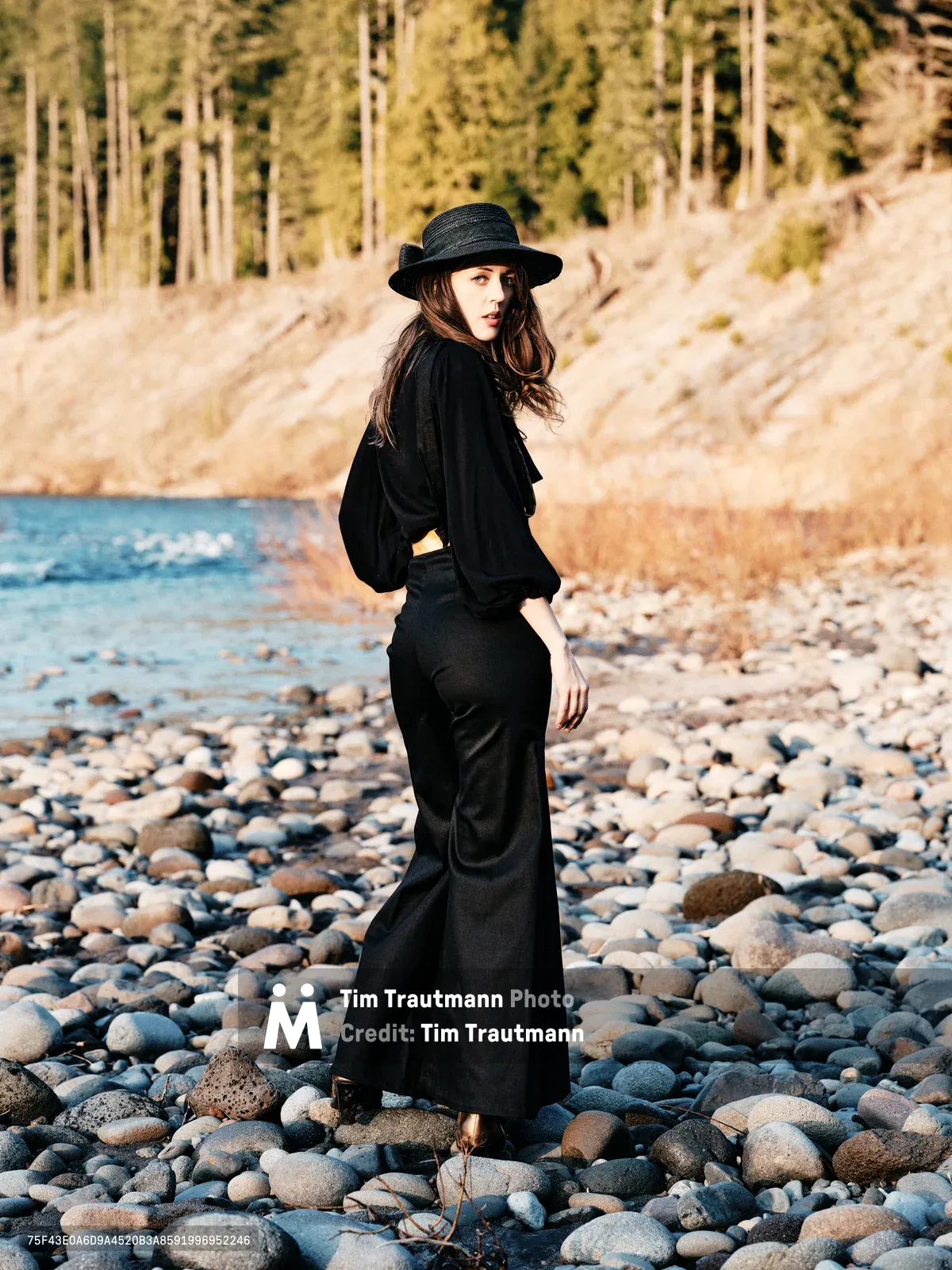 A woman in flowing black attire and wide-brimmed hat stands contemplatively among smooth river stones along the Floodplain Trail in Multnomah County, Oregon. Golden hour light bathes the scene, casting warm tones across the pebbled riverbed while dense forest rises in the soft-focused background. Her pose suggests quiet introspection as she gazes across the water, creating an atmospheric portrait of solitude in nature.