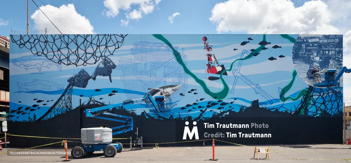 Artists Jeremy Nichols and Devin Finley collaborate on an expansive maritime-themed mural along Southeast 2nd Avenue in Portland's Central Eastside district. The oceanic composition unfolds across a massive wall under partly cloudy skies, featuring flowing kelp, swimming fish, a red and white lighthouse, and dark fishing nets. A blue scissor lift positions one artist at work height while safety barriers cordon off the active creation zone, capturing the monumental scale of Portland Street Art Alliance's community art initiative.