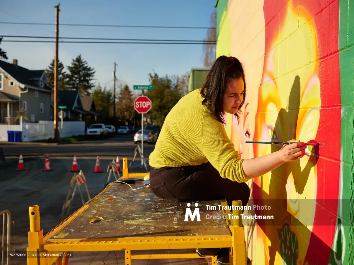 Multidisciplinary artist Villatose works intently on her vibrant Jardín Cubana mural depicting three female figures in Portland's Alberta Arts District, her yellow sweater catching the golden hour light as she applies careful brushstrokes to the colorful wall. The scene captures the intersection of community art and urban revitalization, with residential homes and street infrastructure providing authentic neighborhood context. Her concentrated expression and deliberate movements reflect the meditative focus required for large-scale public art creation.