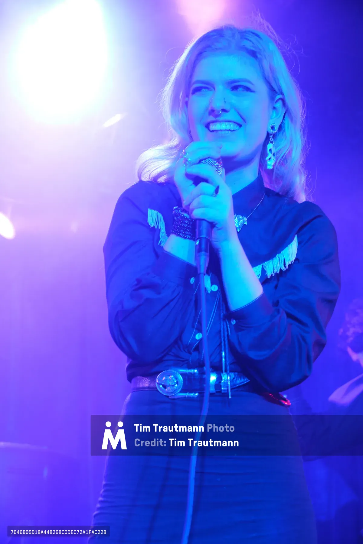 Faith Grossnicklaus of Roselit Bone grips her microphone with both hands, her blonde hair catching the electric blue and purple stage lighting at Dante's club in Portland. The intimate venue's atmospheric lighting transforms the performance space into a dreamy wash of cyan and violet hues, creating an otherworldly backdrop for the violinist's captivating stage presence. Her western-inspired shirt with fringe details and ornate belt buckle blend country aesthetics with the club's moody ambiance.