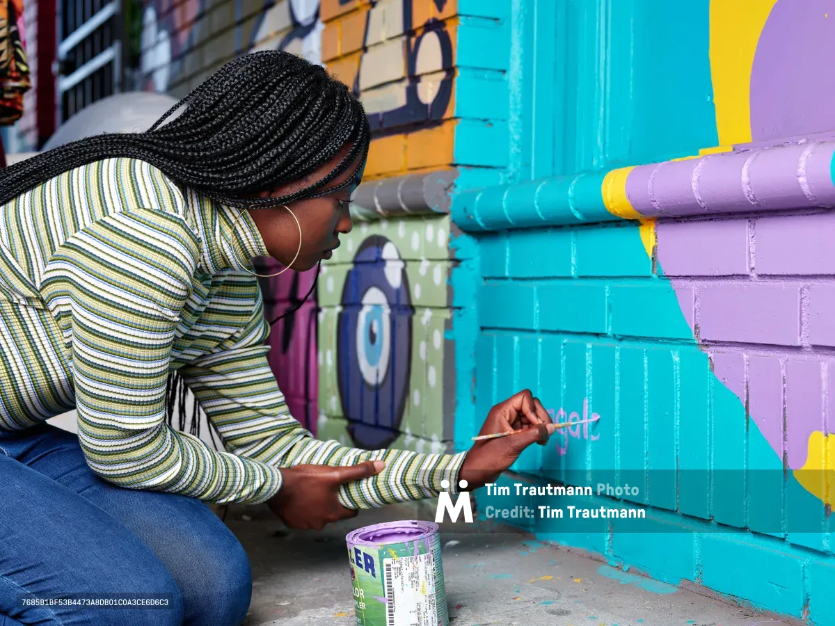 Naomi Likayi, wearing a striped long-sleeve shirt and gold hoop earrings, carefully signs her completed mural on Northeast 15th Avenue in Portland's Alberta Arts District. The artist kneels on the concrete sidewalk, paintbrush in hand, adding her signature to vibrant turquoise and purple geometric shapes that transform the brick wall. Behind her, the colorful street art creates a dynamic backdrop of urban creativity in this historically significant neighborhood.