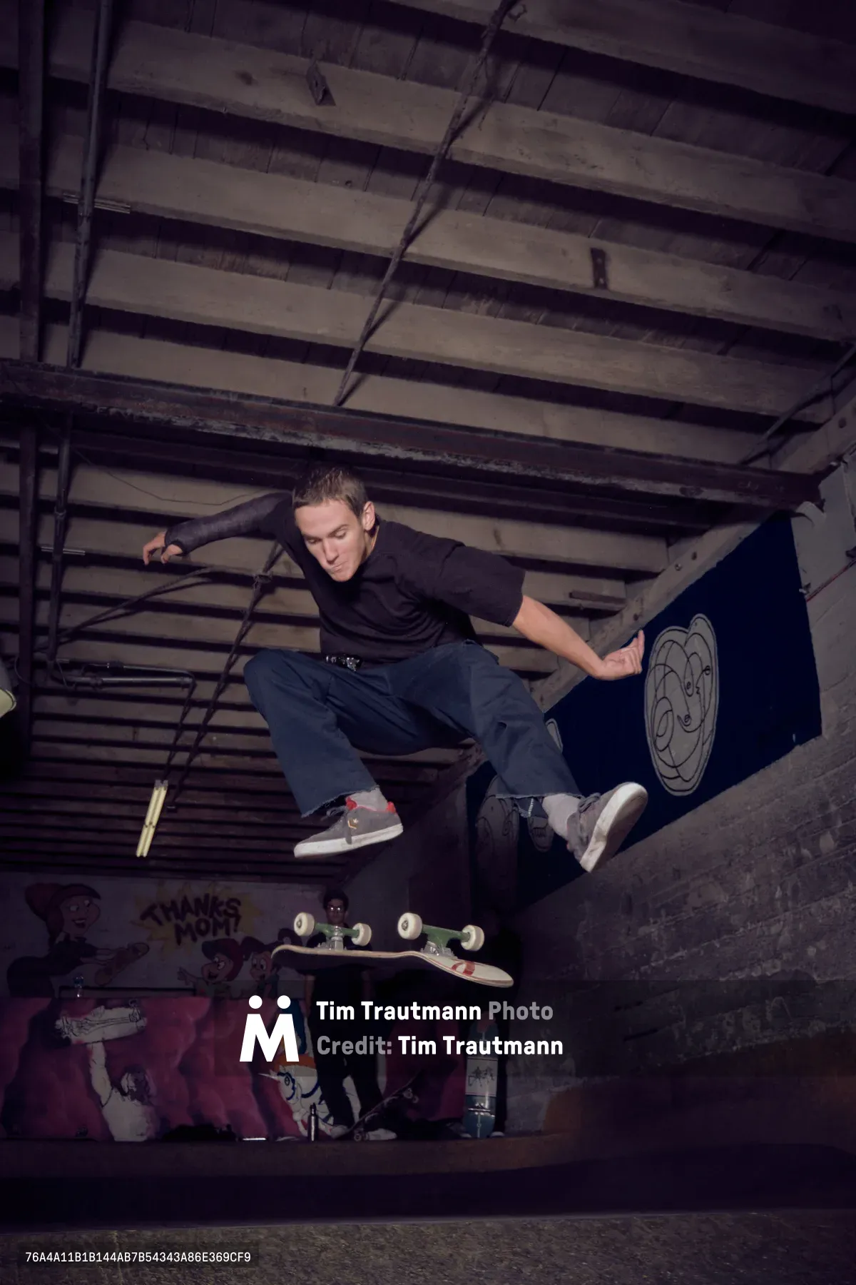 A young skater crouches in focused concentration within the gritty basement confines of Commonwealth Skateboarding in Portland's Buckman neighborhood. The weathered concrete walls and exposed ceiling beams create a moody industrial backdrop, while graffiti and skateboard graphics add splashes of color to the underground space. The atmospheric lighting captures the raw authenticity of Portland's skate culture, with the subject positioned dynamically against the textured urban environment.