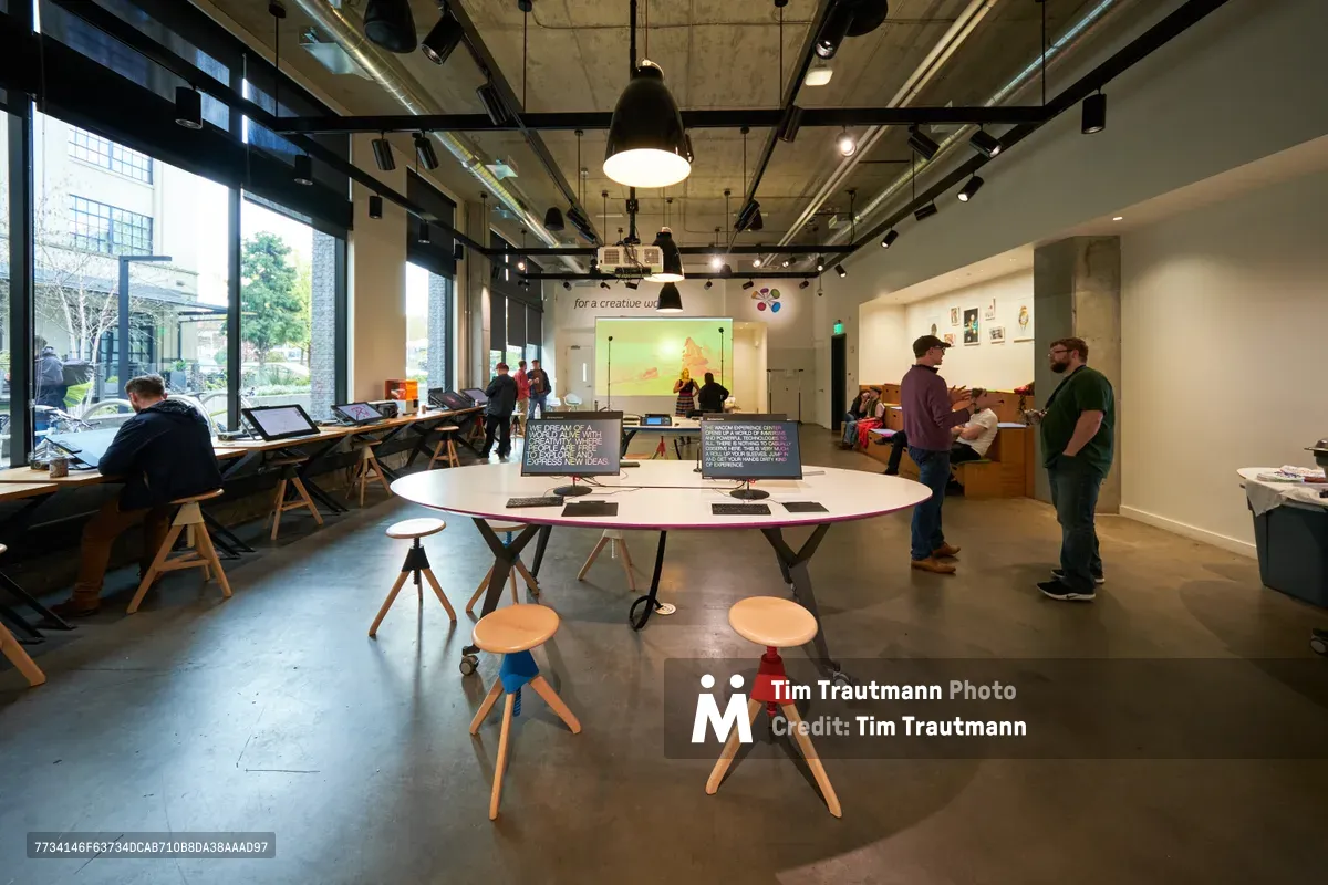 The Wacom Experience Center in Portland's Pearl District buzzes with creative energy as visitors explore digital art workstations and interactive displays. Industrial pendant lights cast warm pools of illumination across polished concrete floors, while floor-to-ceiling windows flood the space with natural daylight from Northwest Irving Street. A central circular table anchored by colorful wooden stools becomes a focal point for collaboration, surrounded by standing workstations where individuals engage with digital drawing tablets and screens displaying artistic content.