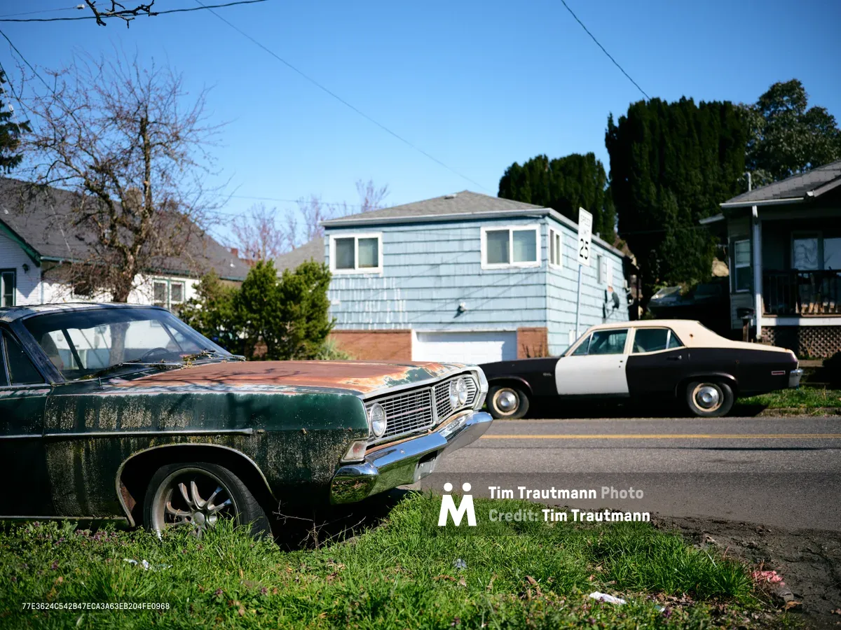 Two weathered American muscle cars from the late 1960s rest along Northeast 15th Avenue near Killingsworth Street in Portland. The foreground features a forest green Chevelle with chrome detailing catching the afternoon light, while a cream-colored classic sits parked across the street. Behind them, modest single-story homes with blue siding and mature evergreen trees create a quintessential Pacific Northwest neighborhood backdrop under clear azure skies.
