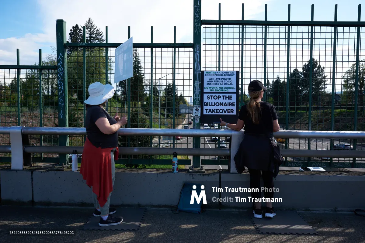Two women stand at a concrete barrier along North Skidmore Street in Portland's Humboldt neighborhood, positioned above Interstate 5 for a May Day banner drop. One activist wears a white sun hat and red skirt, while her companion in dark clothing leans against the railing. Behind them, protest signs reading "STOP THE BILLIONAIRE TAKEOVER" are mounted on green security fencing, creating a scene of grassroots resistance under the Pacific Northwest's characteristic overcast sky.