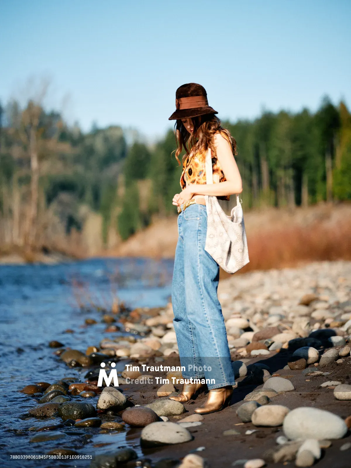 A young woman in vintage-inspired attire stands contemplatively on the rocky shoreline of the Columbia River along Multnomah County's Floodplain Trail. Wearing a distinctive brown felt hat, sunflower-print top, and high-waisted denim, she gazes downward while holding a canvas tote bag. The golden hour light illuminates her figure against the serene water and autumn-touched forest backdrop, creating an intimate moment of reflection in Oregon's natural landscape.