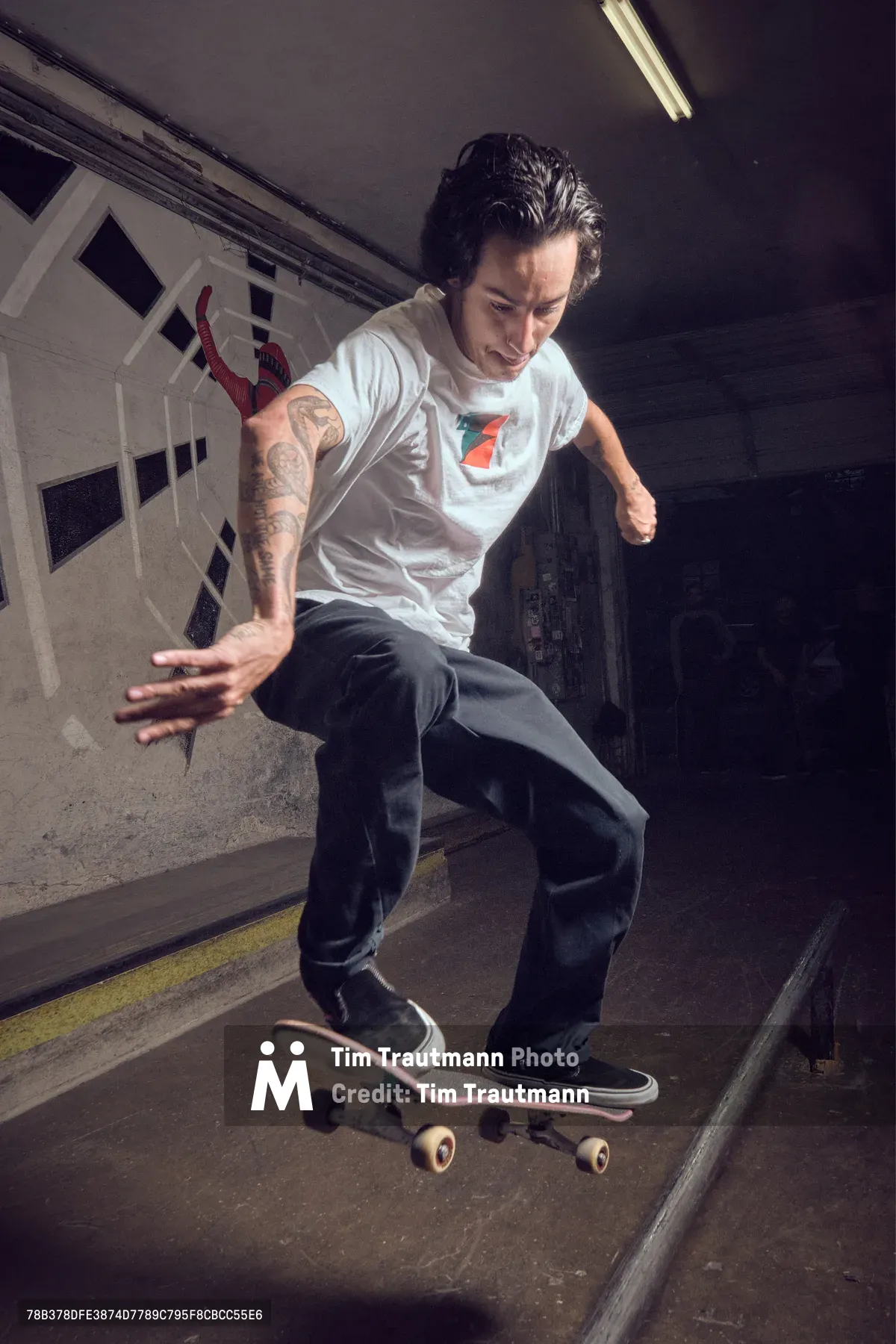 A tattooed skateboarder navigates through the industrial passage of Commonwealth Skateboarding in Portland's Buckman neighborhood. Fluorescent lighting casts stark shadows across concrete walls as the rider demonstrates fluid motion on weathered pavement. The subterranean tunnel creates an intimate urban skating environment, with white structural elements and yellow safety markings defining the confined space.