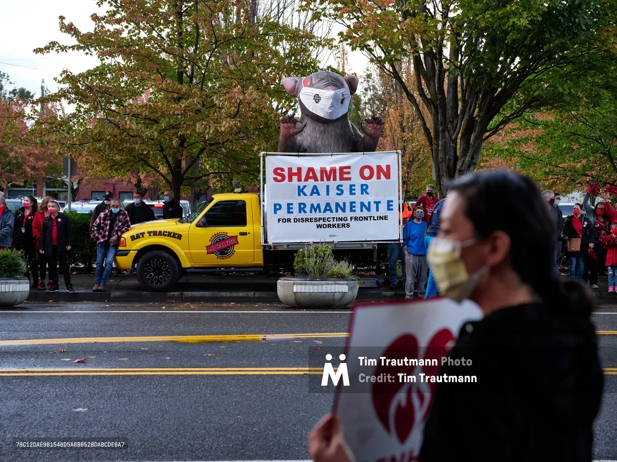 A towering inflatable rat protest figure dominates the scene outside Kaiser Permanente Tower in Portland's Lloyd District, its menacing presence amplified by autumn's golden canopy. Red-clad nurses and supporters gather in solidarity along Northeast Multnomah Street, their masked faces reflecting both pandemic precautions and labor tensions. The bold white protest sign declaring 'SHAME ON KAISER PERMANENTE' cuts through the overcast afternoon light, while a yellow Rat Tracker truck anchors the demonstration with industrial determination.