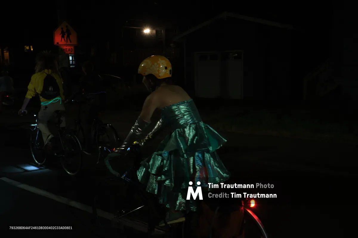 Under the cover of Portland's Richmond neighborhood darkness, two cyclists navigate the streets in an enchanting juxtaposition of formal elegance and urban mobility. The foreground rider wears a lustrous teal gown that catches the sparse streetlight, creating ethereal ripples of fabric against the night. A golden helmet provides a striking contrast to the flowing dress, while warning signs and distant street lamps punctuate the mysterious nocturnal scene.