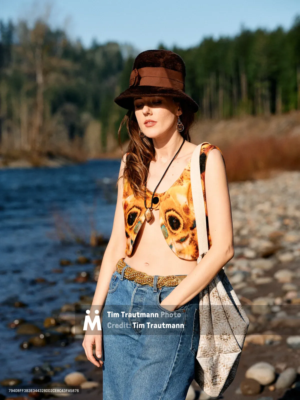 A young woman poses confidently along Oregon's Floodplain Trail, wearing an eye-catching owl-print tank top paired with high-waisted denim and a dark felt hat. The golden hour light illuminates her relaxed stance against the backdrop of the Columbia River's shimmering waters and the verdant Pacific Northwest forest. Her eclectic accessories, including layered necklaces and a woven bag, complete this portrait of contemporary bohemian style in a natural riverside setting.