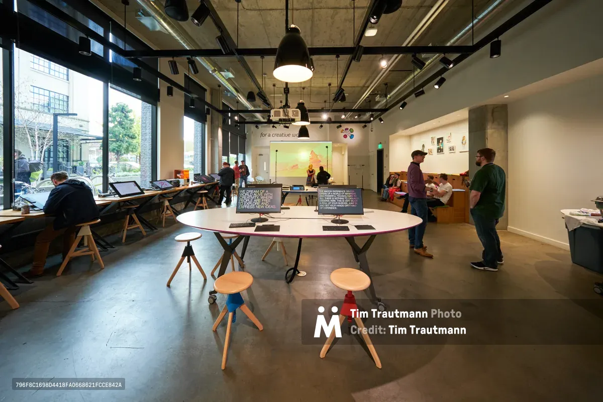 The bustling interior of Portland's Wacom Experience Center reveals a vibrant creative workspace where artists and designers gather to explore digital artistry. Floor-to-ceiling windows flood the industrial-chic space with natural light, illuminating collaborative workstations equipped with drawing tablets and monitors. Small groups of creators engage in hands-on learning around sculptural white tables, while others work intently at individual stations lined along the exposed concrete walls. The open-plan design, punctuated by pendant lighting and polished concrete floors, embodies Portland's innovative spirit in the heart of the Pearl District.