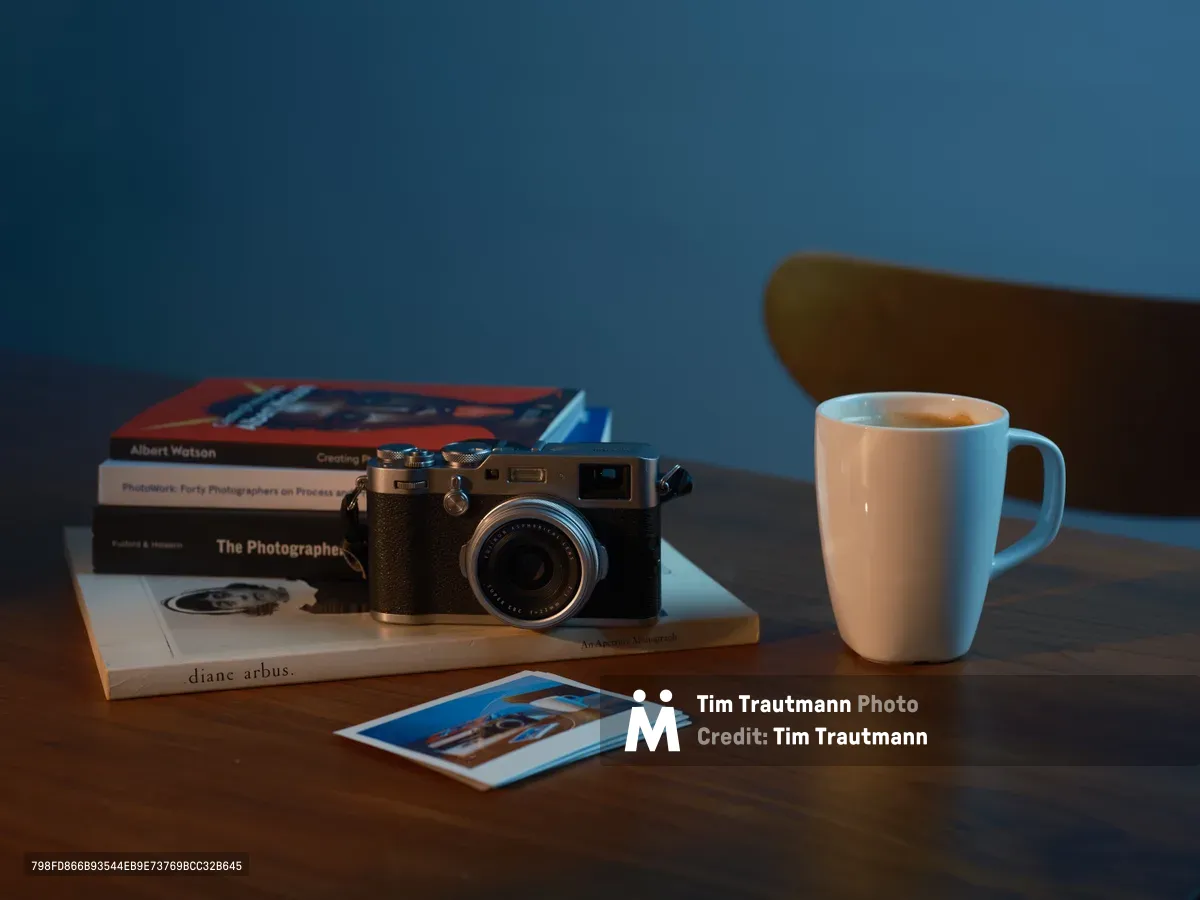 Golden hour light streams across a wooden desk in a Richmond District home, illuminating the tools of a contemplative photographer. A vintage rangefinder camera rests atop a carefully curated stack of photography books including works by Albert Watson and Diane Arbus, while a white ceramic mug holds steaming coffee nearby. Scattered color photographs lie casually beside the stack, suggesting a morning ritual of inspiration and caffeine in this Portland creative space.