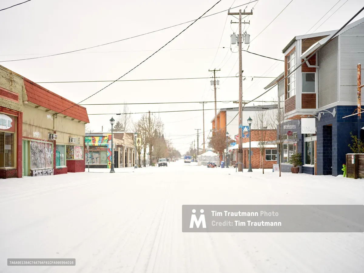 A blanket of pristine snow transforms Northeast Alberta Street in Portland's Alberta Arts District into a hushed winter tableau. The eclectic mix of colorful storefronts - from the mint green corner shop to modern mixed-use buildings - stands muted against the overcast sky, while power lines create geometric patterns overhead. Bare trees punctuate the scene as a lone vehicle navigates the snow-covered street, capturing the quiet beauty of an urban neighborhood in winter's grip.