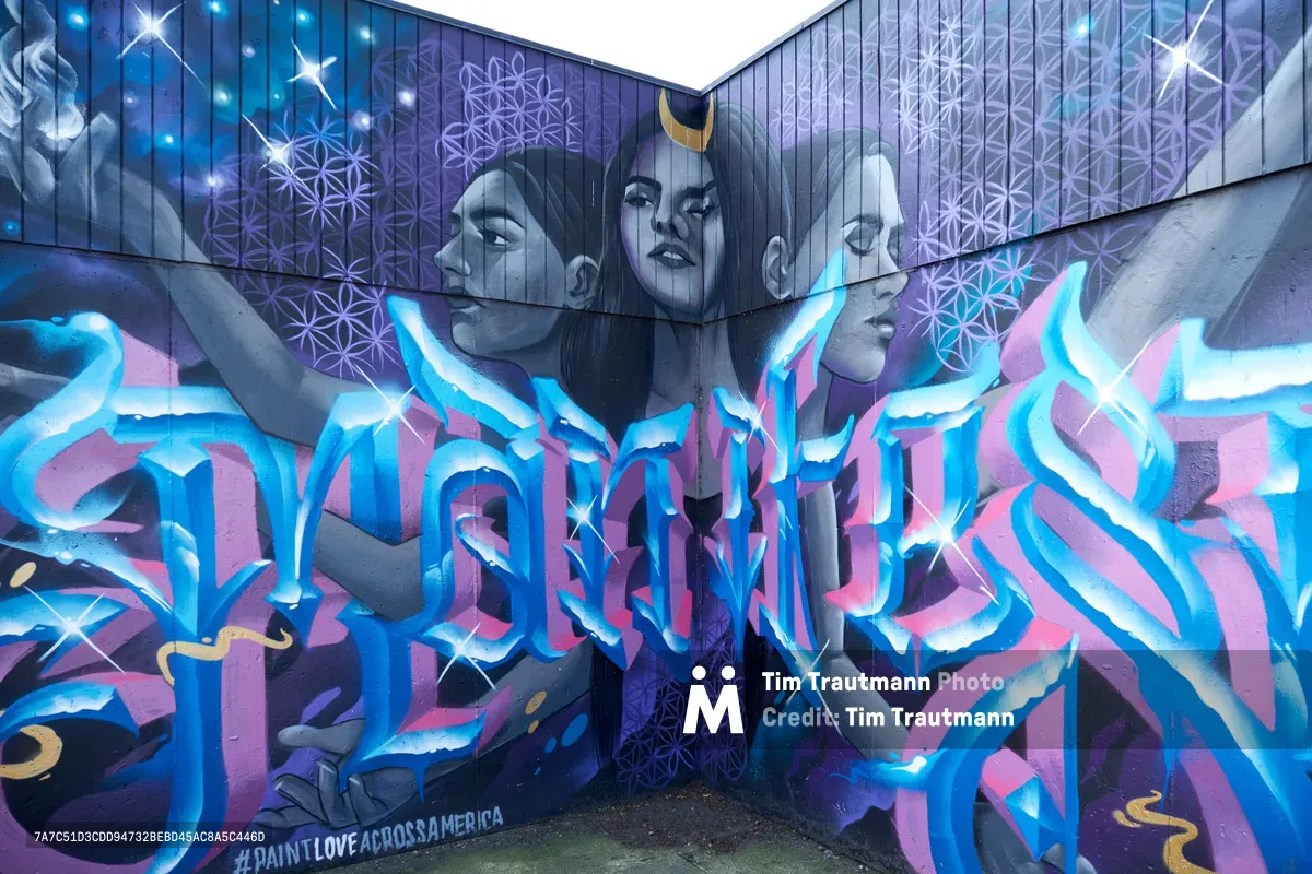 Three ethereal faces crowned with golden crescents gaze from a corner mural in Portland's Central Eastside, their monochromatic features floating above an explosive wildstyle composition in electric blues and magentas. Sacred geometry patterns of interconnected circles shimmer across the upper walls like constellation maps, while dimensional letterforms surge below in fluid, ribbon-like strokes that seem to dance with otherworldly energy. The #PAINTLOVEACROSSAMERICA hashtag anchors this transcendent piece where street art meets spiritual iconography.